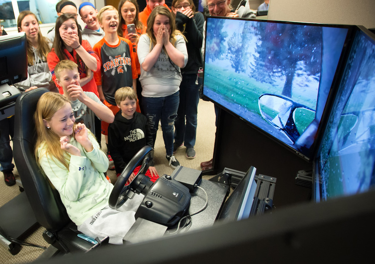 A young girl drives a driving simulator. Her peers around her look surprised.