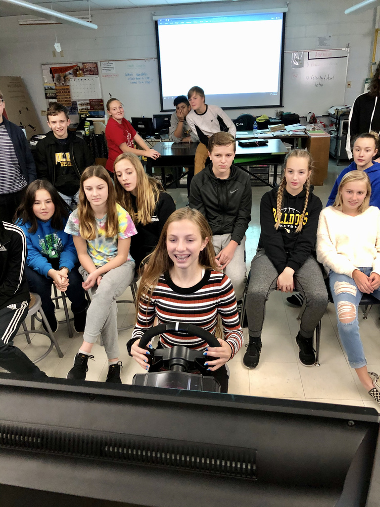 A group of students watch a classmate drive a driving simulator. 